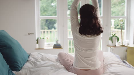 Rear-view-of-mature-Asian-woman-in-pyjamas-sitting-on-bed-meditating-in-yoga-pose---shot-in-slow-motion