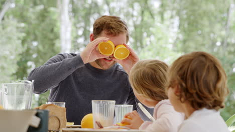 Vater-Bringt-Kinder-Am-Frühstückstisch-Zum-Lachen,-Indem-Er-Gesichter-Mit-Geschnittenen-Orangen-Als-Augen-Formt