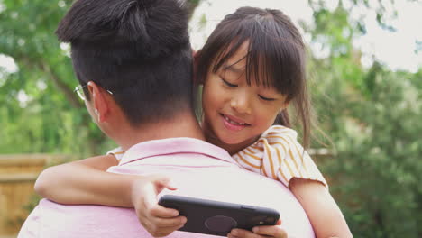 Asiatischer-Vater-Kuschelt-Mädchen-Im-Garten,-Während-Das-Mädchen-über-Seine-Schulter-Auf-Sein-Mobiltelefon-Schaut