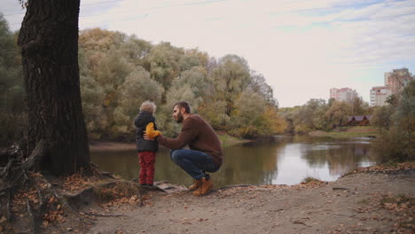 family-rest-in-autumn-forest-father-is-speaking-to-child-son-and-showing-picturesque-lake-in-forest-hiking-at-nature-at-autumn-weekend