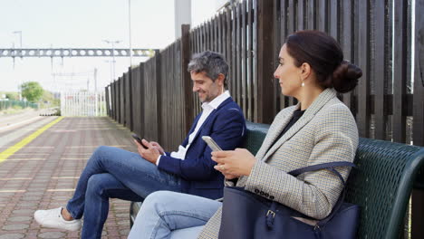 Business-Commuters-Sitting-On-Seat-On-Railway-Platform-With-Mobile-Phones