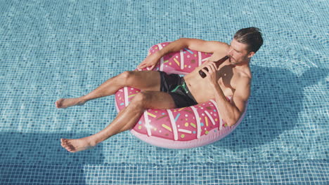 Overhead-Shot-Of-Man-In-Swim-Shorts-Floating-On-Inflatable-Ring-In-Swimming-Pool-Drinking-Beer