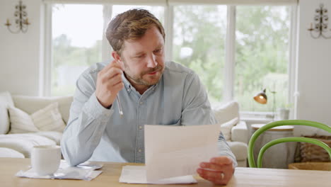 Worried-Man-Sitting-At-Table-At-Home-Reviewing-Domestic-Finances-Looking-At-Bill