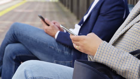 Close-Up-Of-Business-Commuters-Sitting-On-Seat-On-Railway-Platform-With-Mobile-Phones