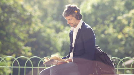 Hombre-De-Negocios-Leyendo-Un-Libro-Y-Escuchando-Con-Auriculares-En-El-Parque-Con-Café-Para-Llevar