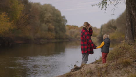 woman-and-little-child-are-standing-on-coast-of-river-at-autumn-day-family-weekend-at-nature-mother-and-son-are-walking-in-forest-calm-and-happy-time-together