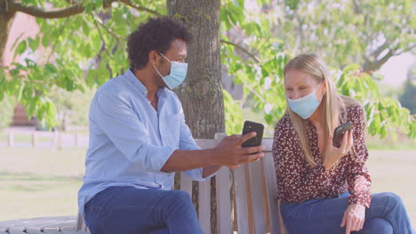 Una-Pareja-Socialmente-Distanciada-Con-Máscaras-Se-Reúne-En-El-Parque-Durante-Una-Pandemia-De-Salud-Mirando-Teléfonos-Móviles