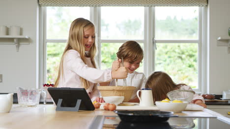 Children-In-Pyjamas-Making-Pancakes-In-Kitchen-At-Home-Following-Recipe-On-Digital-Tablet