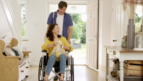 Mature-Asian-man-pushing-wife-in-wheelchair-in-hallway-at-home-returning-from-shopping-trip-holding-reusable-bag---shot-in-slow-motion