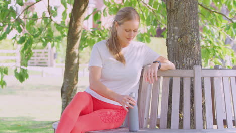 Woman-Wearing-Fitness-Clothing-Sitting-On-Seat-Under-Tree-Checking-Activity-Monitor-On-Smartwatch