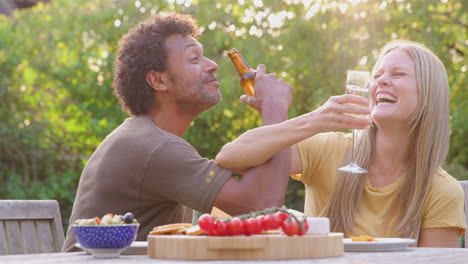 Mature-Couple-Link-Arms-As-They-Celebrate-With-Champagne-And-Beer-At-Table-In-Garden-With-Snacks