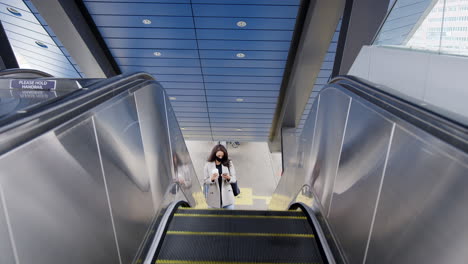 Businesswoman-On-Escalator-At-Railway-Station-With-Mobile-Phone-Wearing-PPE-Face-Mask-In-Pandemic