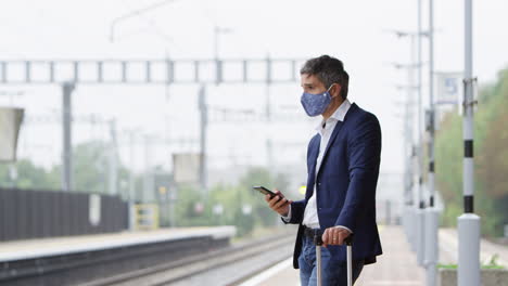 Businessman-On-Railway-Platform-With-Mobile-Phone-Wearing-PPE-Face-Mask-During-Health-Pandemic