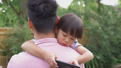 Asian-Father-Cuddling-Son-In-Garden-As-Boy-Looks-Over-His-Shoulder-At-Mobile-Phone