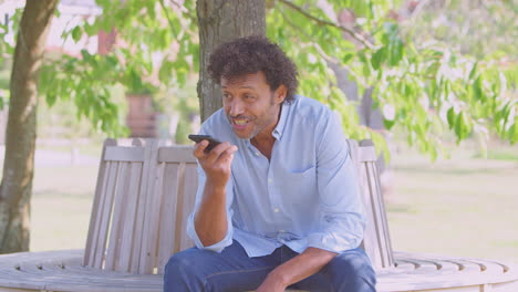 Mature-Man-Sitting-On-Park-Bench-Under-Tree-In-Summer-Talking-On-Mobile-Phone-Using-Microphone