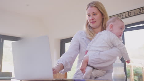 Working-mother-using-laptop-at-home-whilst-cuddling-baby-son-over-shoulder--shot-in-slow-motion