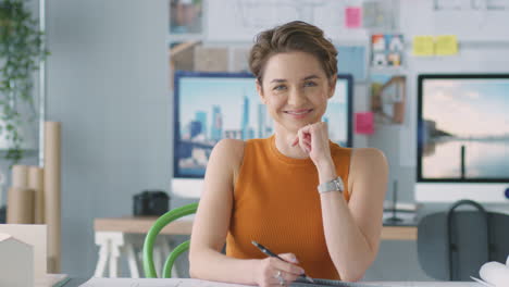 Portrait-Of-Smiling-Female-Architect-In-Office-Working-At-Desk-With-Model-Of-Building