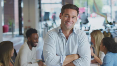 Portrait-Of-Businessman-With-Colleagues-In-Background-Sitting-Around-Table-In-Open-Plan-Office
