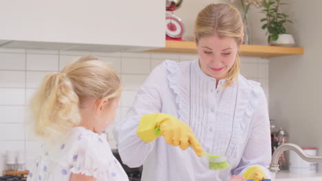 Mother-wearing-rubber-gloves-at-home-in-kitchen-with-young-daughter-having-fun-as-girl-puts-feet-in-sink-as-they-do-washing-up-at-sink--shot-in-slow-motion
