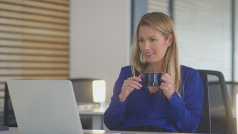 Young-Businesswoman-With-Hot-Drink-Working-Late-Sitting-At-Desk-With-Laptop-In-Open-Plan-Office