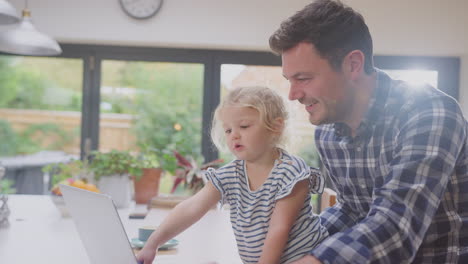 Berufstätiger-Vater-Benutzt-Laptop-Zu-Hause-Auf-Der-Küchentheke,-Während-Er-Sich-Um-Seine-Kleine-Tochter-Kümmert-–-Aufnahme-In-Zeitlupe