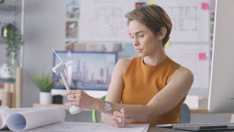 Female-Architect-In-Office-Working-On-Drawings-For-Renewable-Energy-Project-With-Model-Wind-Turbine