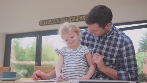 Father-at-home-at-kitchen-counter-helping-young-daughter-to-draw-picture-in-book---shot-in-slow-motion