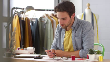 Male-Owner-Of-Fashion-Business-Working-On-Designs-Receives-Message-On-Mobile-Phone-Sitting-At-Desk