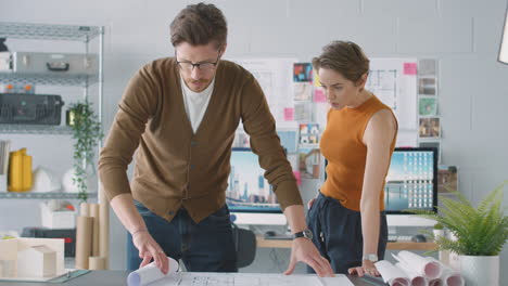 Male-And-Female-Architects-In-Office-Unrolling-Plan-Or-Blueprint-And-Laying-It-On-Desk