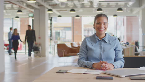 Portrait-Of-Businesswoman-Working-At-Desk-In-Modern-Open-Plan-Office-With-Colleagues-In-Background
