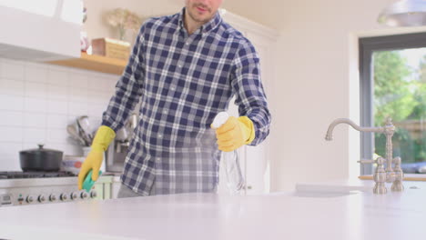 Man-at-home-in-kitchen-wearing-rubber-gloves-cleaning-down-work-surface-using-cleaning-spray---shot-in-slow-motion