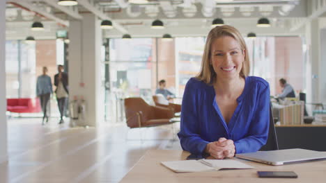 Portrait-Of-Businesswoman-Working-At-Desk-In-Modern-Open-Plan-Office-With-Colleagues-In-Background