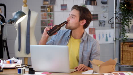 Male-Owner-Of-Fashion-Business-With-Bottle-Of-Beer-Listening-To-Music-On-Laptop-In-Studio