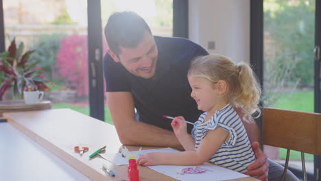 Father-and-young-daughter-having-fun-at-home-sitting-at-table-and-painting-decoration-together---shot-in-slow-motion