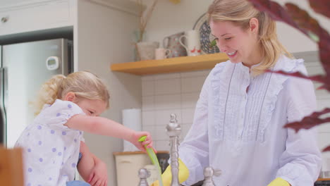 Mother-wearing-rubber-gloves-at-home-in-kitchen-with-young-daughter-having-fun-as-they-do-washing-up-at-sink--shot-in-slow-motion