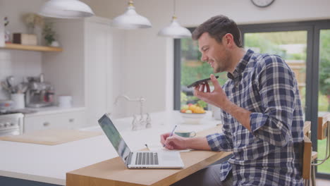 Man-working-from-home-using-laptop-on-kitchen-counter-talking-into-microphone-of-mobile-phone--shot-in-slow-motion