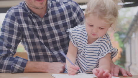 Father-at-home-at-kitchen-counter-helping-young-daughter-to-draw-picture-in-book---shot-in-slow-motion