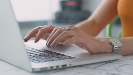Close-Up-Showing-Hands-Of-Female-Architect-In-Office-Working-At-Desk-On-Laptop-With-Plans