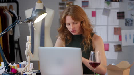 Female-Owner-Of-Fashion-Business-With-Glass-of-Wine-Working-Late-On-Laptop-In-Studio