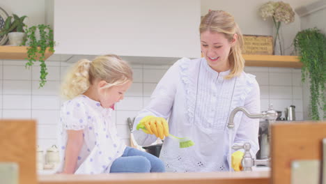 Mother-wearing-rubber-gloves-at-home-in-kitchen-with-young-daughter-having-fun-as-girl-puts-feet-in-sink-as-they-do-washing-up-at-sink--shot-in-slow-motion
