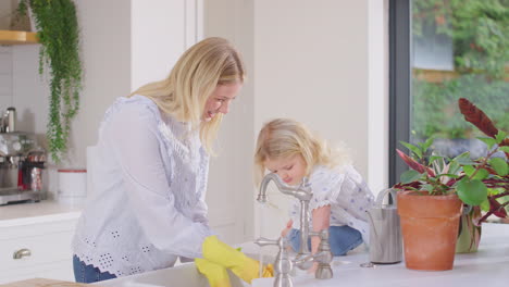 Panning-shot-of-mother-at-home-in-kitchen-with-young-daughter-wearing-rubber-gloves-and-doing-washing-up-at-sink--shot-in-slow-motion