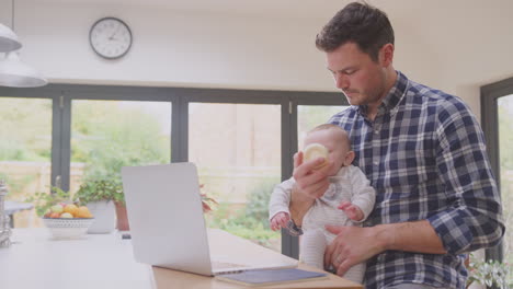 Working-father-using-laptop-at-home-whilst-feeding-baby-son-sitting-on-his-knee--from-bottle---shot-in-slow-motion