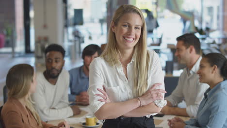 Portrait-Of-Businesswoman-With-Colleagues-In-Background-Sitting-Around-Table-In-Open-Plan-Office