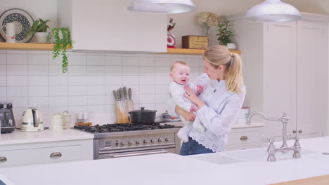 Loving-mother-carrying-laughing-baby-son-around-kitchen-at-home-playing-game-together