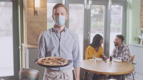 Porträt-Eines-Kellners-Im-Restaurant-Mit-Maske,-Der-Einem-Paar-Während-Einer-Gesundheitspandemie-Pizza-Serviert