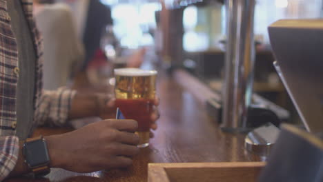 Close-Up-Of-Male-Customer-In-Bar-Making-Contactless-Payment-For-Drinks-In-Health-Pandemic