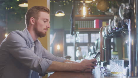 Unhappy-Man-Sitting-At-Pub-Bar-Drinking-Alone-With-Glass-Of-Whisky-Sending-Message-On-Mobile-Phone