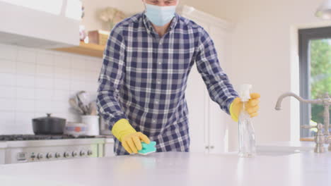 Mann-Mit-Gesichtsmaske-Zu-Hause-In-Der-Küche,-Trägt-Gummihandschuhe-Und-Reinigt-Die-Arbeitsfläche-Mit-Reinigungsspray-Während-Einer-Gesundheitspandemie-–-Aufgenommen-In-Zeitlupe