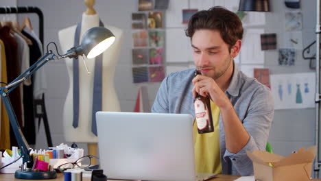 Smiling-Male-Owner-Of-Fashion-Business-With-Bottle-Of-Beer-Working-Late-On-Laptop-In-Studio