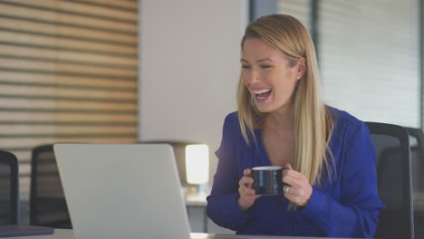 Young-Businesswoman-With-Hot-Drink-Having-Late-Video-Chat-On-Laptop-Working-In-Open-Plan-Office